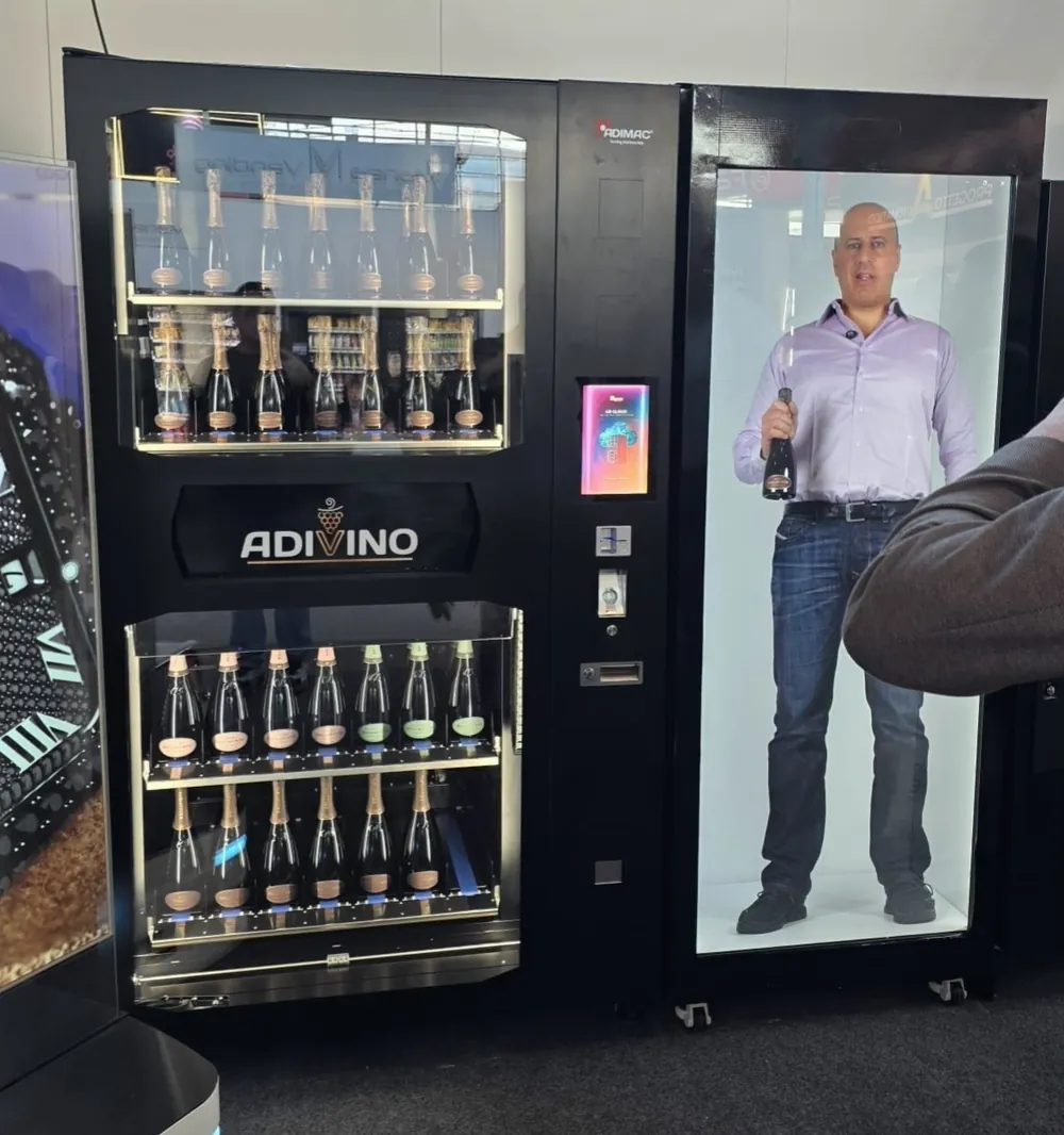 At a trade show, a life-size holographic presenter holding a bottle stands in a display cabinet next to a wine vending machine.
